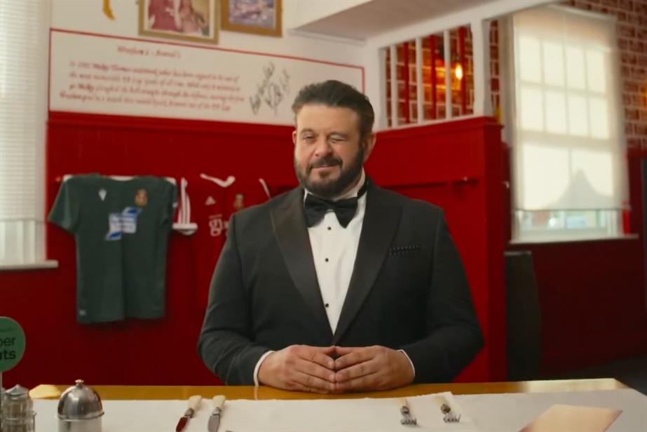 Adam Richman wears a bow tie and suit. Sitting down at a dining table, he winks at the camera.