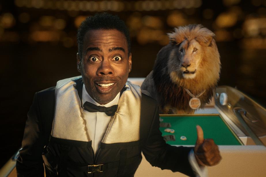 Chris Rock speaks to camera with a lion sitting at a poker table in the background.
